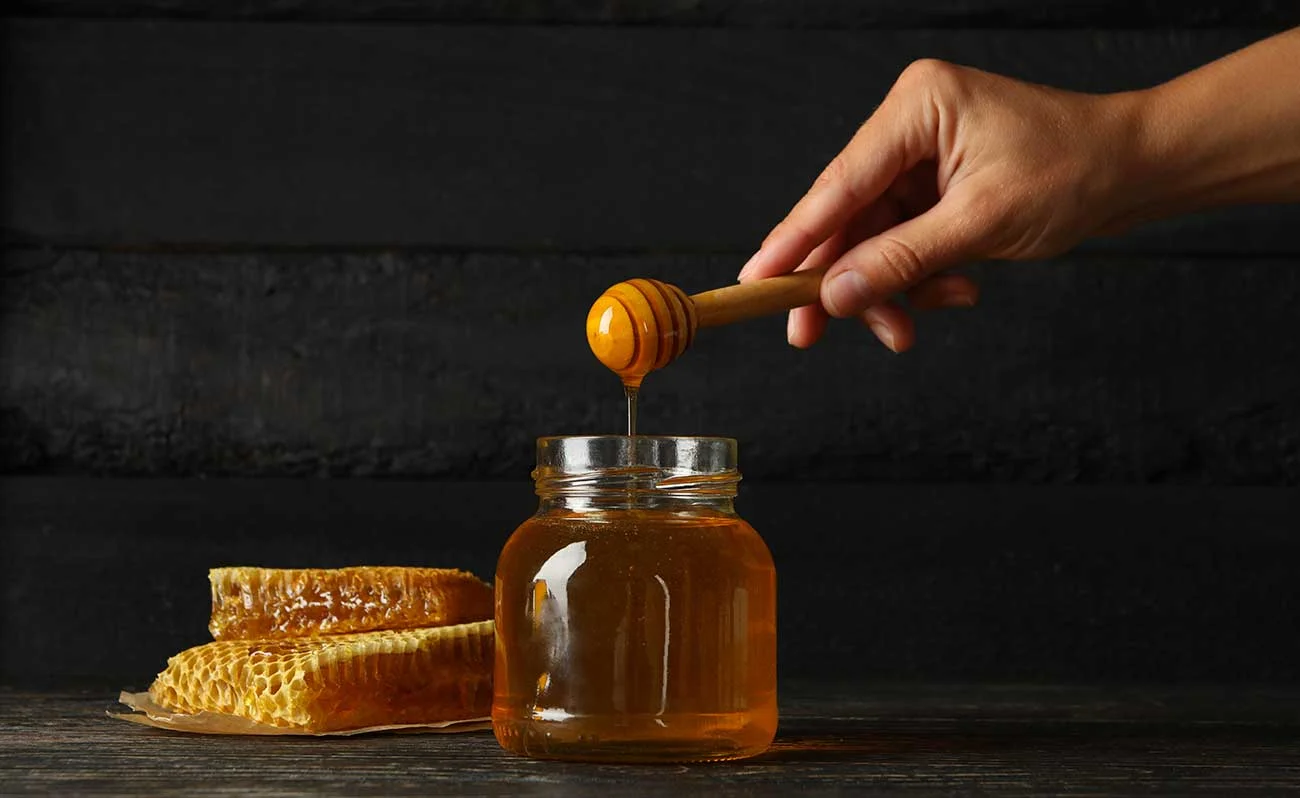 Female hand holds dipper with honey on wooden background with jar and honeycombs