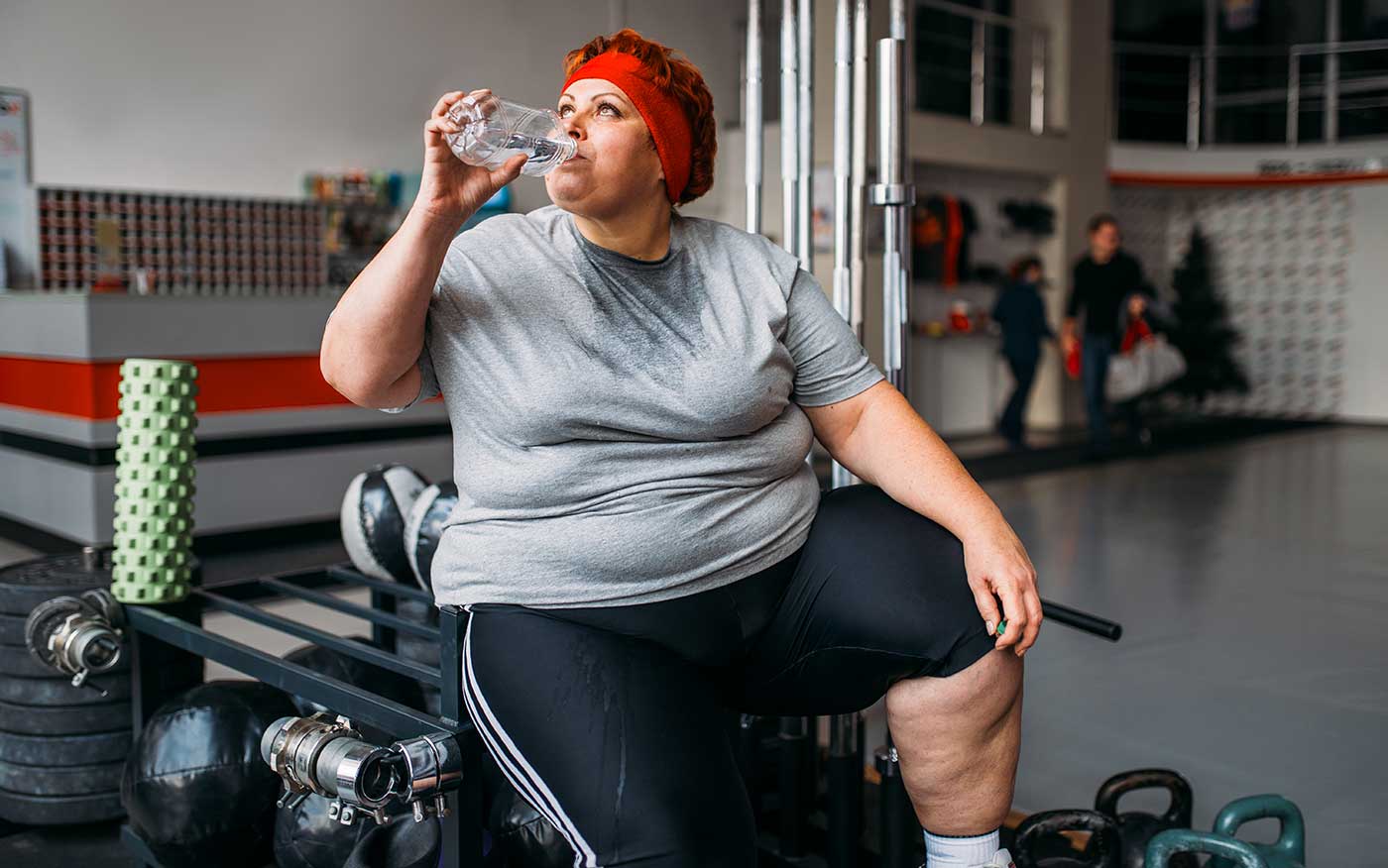 Fat woman drinks water after training in gym